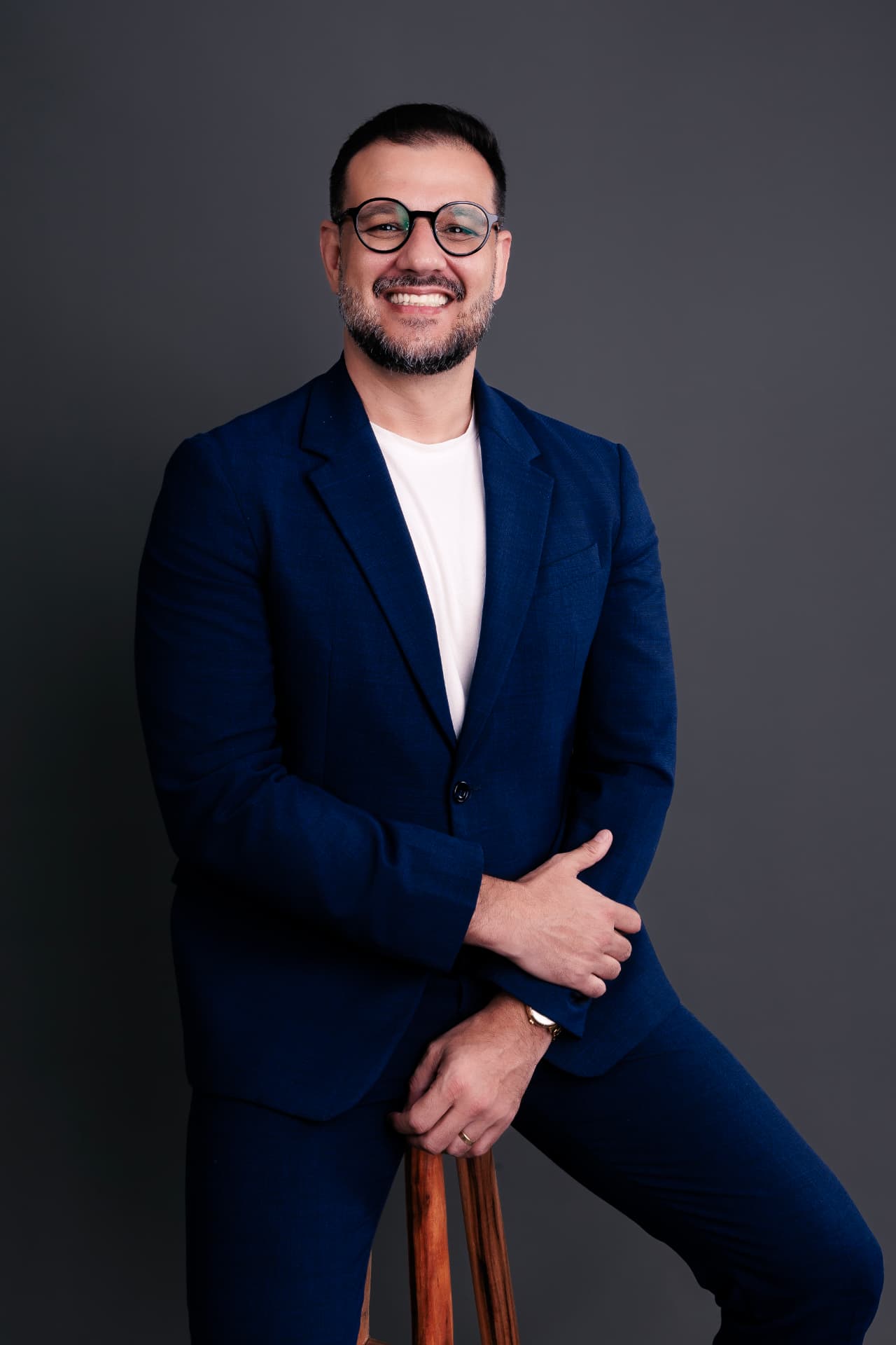 Leandro Albuquerque sorrindo, de blazer azul-marinho e camiseta branca, apoiado em um banco alto